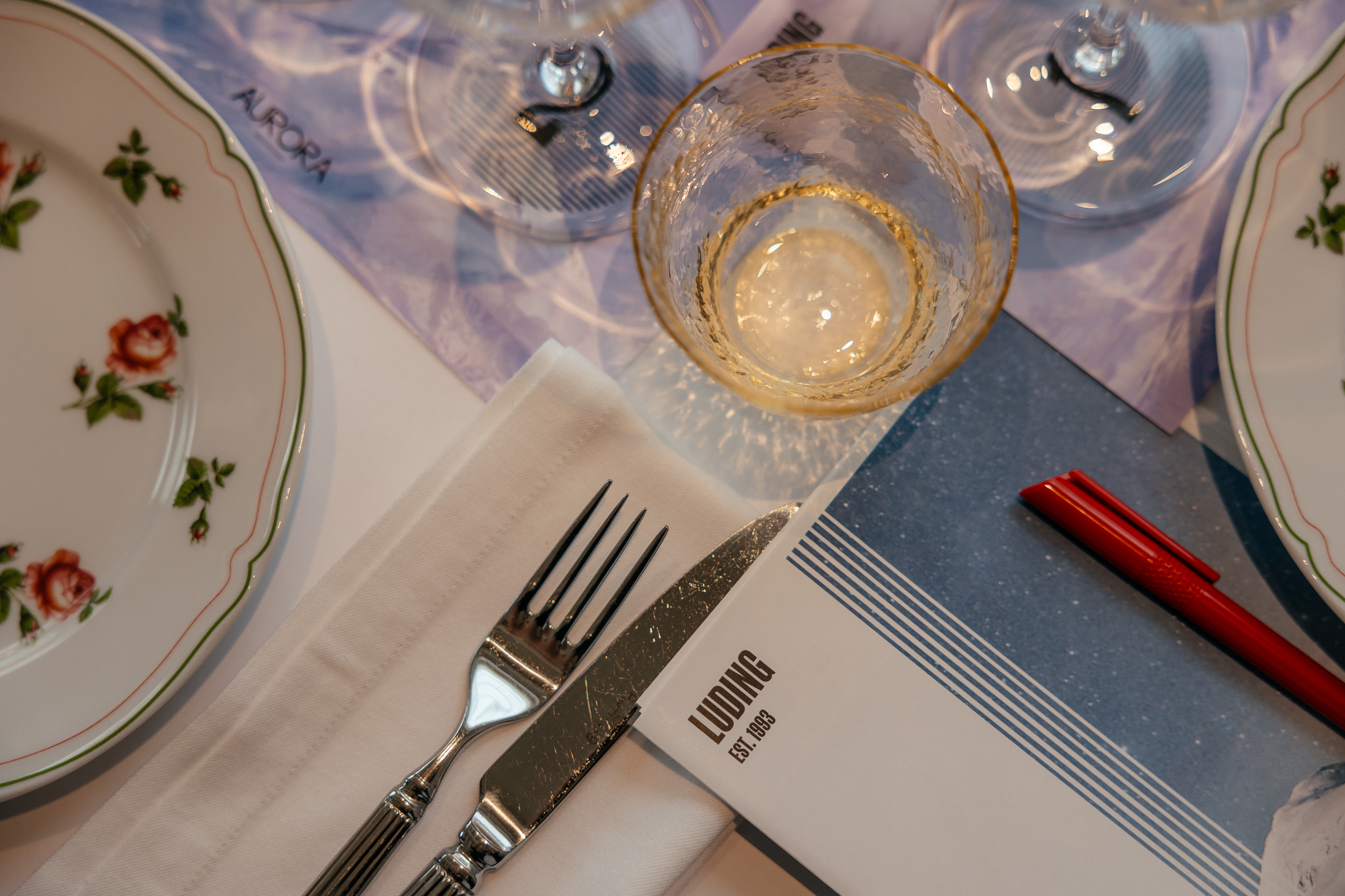 Красная ручка и буклет с логотипом LUDING (ЛУДИНГ) на столе рядом с бокалами, тарелкой и столовыми приборами на белой салфетке